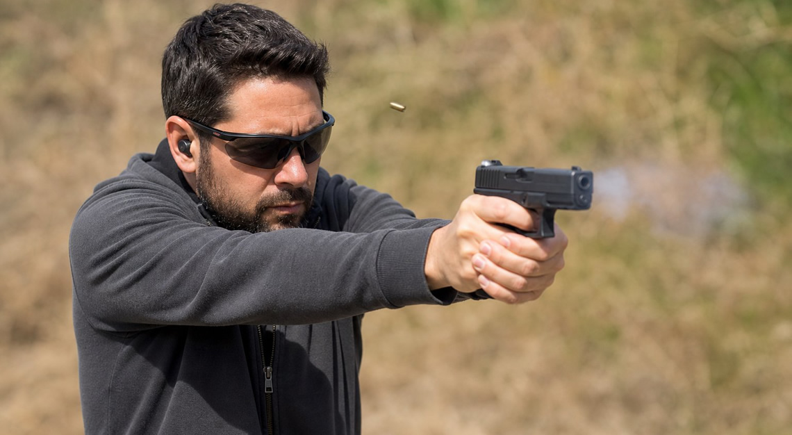 An adult aiming a handgun with both hands in a self-defense shooting stance outdoors at a shooting range.